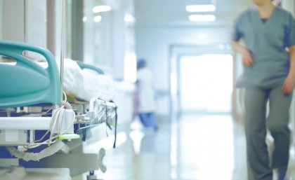 A hospital corridor with a trolley bed on one side, and an out of focus outline of a hospital worker in green scrubs on the other. 